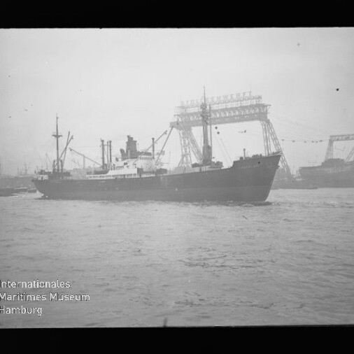 Schwarzweißfotografie eines Frachtschiffs, das in Hafennähe über leicht bewegte See fährt, vor großen Werftkranen und einer hohen Portalkrankonstruktion mit Girlanden, links unten das Logo des Internationalen Maritimen Museums Hamburg.
