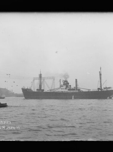 Schwarzweiß-Foto eines großen Dampfschiffs mittig auf dem Wasser, mit Rauch aus dem Schornstein, im Vordergrund wellige Wasserfläche und links ein kleines Boot, im Hintergrund Hafenkräne und Industrieanlagen; im unteren linken Bildbereich das Logo des Internationalen Maritimen Museums Hamburg.