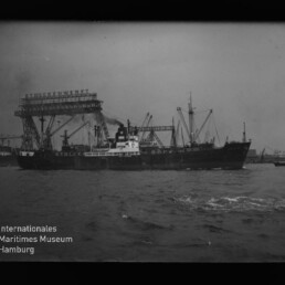 Schwarzweißfoto eines Frachtschiffs mit rauchendem Schornstein, das im Hafen vor hohen Krananlagen und einer Förderbrücke liegt; wellige Wasseroberfläche im Vordergrund und das Logo des Internationalen Maritimen Museums Hamburg unten links.