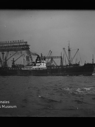 Schwarzweißfoto eines Frachtschiffs mit rauchendem Schornstein, das im Hafen vor hohen Krananlagen und einer Förderbrücke liegt; wellige Wasseroberfläche im Vordergrund und das Logo des Internationalen Maritimen Museums Hamburg unten links.