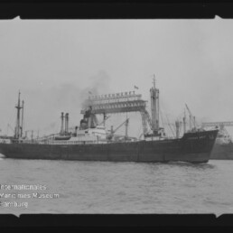 Schwarzweißaufnahme eines Frachtschiffs, das auf dem Wasser an einer Werft mit großen Auslegerkränen und dem Schriftzug „Stülckenwerft“ vorbeifährt; unten links ist ein Museumswasserzeichen zu sehen und der Filmrand bleibt erkennbar.