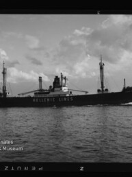 Schwarzweißaufnahme eines Frachtschiffs der Reederei Hellenic Lines mit sichtbarem Schriftzug auf dem Rumpf, Ladebäumen und Schornstein, das bei ruhiger See unter teilweise bewölktem Himmel von links nach rechts fährt; unten links das Logo des Internationalen Maritimen Museums Hamburg.