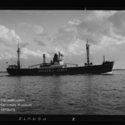 Schwarzweißaufnahme eines Frachtschiffs der Reederei Hellenic Lines mit sichtbarem Schriftzug auf dem Rumpf, Ladebäumen und Schornstein, das bei ruhiger See unter teilweise bewölktem Himmel von links nach rechts fährt; unten links das Logo des Internationalen Maritimen Museums Hamburg.