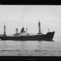 Schwarzweiß-Fotografie eines Frachtschiffs in Seitenansicht auf ruhiger See, mit dunklem Rumpf, mittiger Aufbauten und Schornstein sowie mehreren Ladekranen; unten links sichtbares Wasserzeichen des Internationalen Maritimen Museums Hamburg.