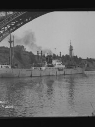 Schwarzweißaufnahme eines Dampfschiffs, das unter einer großen stählernen Bogenbrücke auf einem ruhigen Fluss fährt, mit Rauch aus dem Schornstein, Uferböschung im Hintergrund und Spiegelung im Wasser; Logo des Internationalen Maritimen Museums Hamburg im Bild.