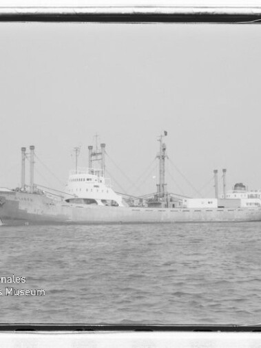 Schwarzweiß-Fotografie eines Frachtschiffs in ruhigem Wasser, seitliche Ansicht mit mehreren Masten und Ladebäumen, Industrieanlagen am Ufer im Hintergrund und Museumslogo „Internationales Maritimes Museum Hamburg“ unten links.