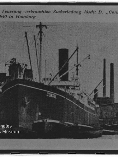 Schwarzweiß-Foto eines großen Frachtschiffs mit der Aufschrift ‚CONSUL‘ an der Hafenmole, flankiert von Schleppern, vor Hafenkranen und hohen Schornsteinen; oben eine deutsche Bildunterschrift und unten links das Logo des Internationalen Maritimen Museums Hamburg.
