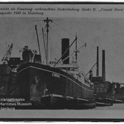 Schwarzweiß-Foto eines großen Frachtschiffs mit der Aufschrift ‚CONSUL‘ an der Hafenmole, flankiert von Schleppern, vor Hafenkranen und hohen Schornsteinen; oben eine deutsche Bildunterschrift und unten links das Logo des Internationalen Maritimen Museums Hamburg.