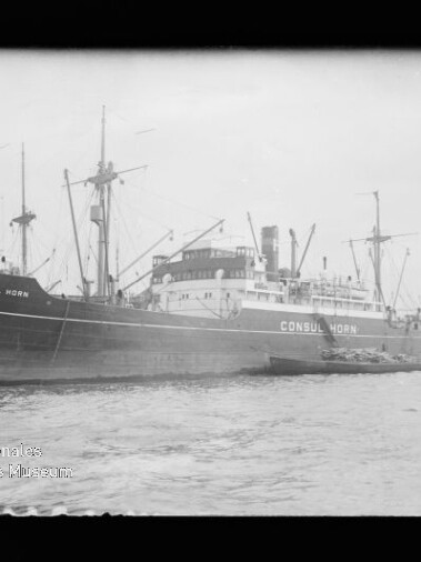 Schwarzweiß-Foto eines großen Frachtschiffs mit dem Namen CONSUL HORN, das am Kai liegt; mehrere Masten und ein Schornstein sind sichtbar, kleinere Boote liegen am Rumpf und Wasserzeichen des Internationalen Maritimen Museums Hamburg ist unten links zu sehen.