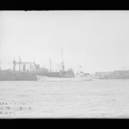 Schwarzweißaufnahme eines Hafens mit einem weißen Passagier- oder Dienstboot im Vordergrund vor einem großen, am Kai liegenden Frachtschiff mit der Aufschrift DOCK VII und mehreren Hafenkränen im Hintergrund.