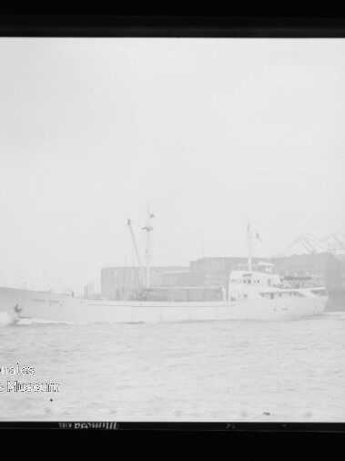 Weißes Frachtschiff mit Aufbauten und zwei Masten in einem nebligen Industriehafen; im Hintergrund Lagergebäude und mehrere Hafenkräne, im Vordergrund wellige Wasseroberfläche und unten links ein Museumsstempel.