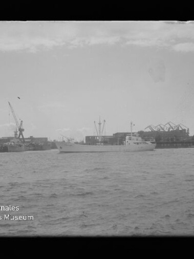 Schwarzweißaufnahme eines mittleren weißen Schiffes auf dem Wasser vor einer Werft mit Kränen und Hallen, mit Museumslogo unten links.