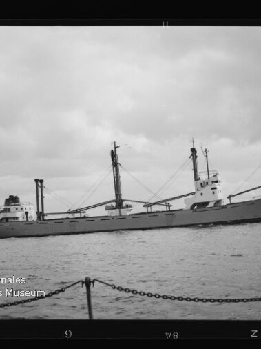 Schwarzweißfoto eines Frachtschiffs mit drei Ladekränen und langem Rumpf auf ruhiger See unter wolkenverhangenem Himmel; im Vordergrund Ketten eines Kais und links unten das Logo des Internationalen Maritimen Museums Hamburg.