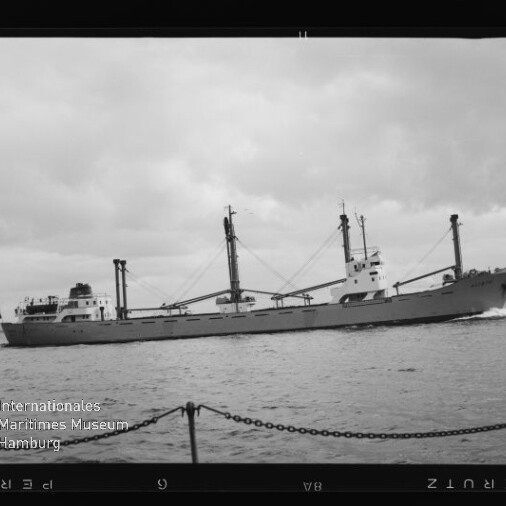 Schwarzweißfoto eines Frachtschiffs mit drei Ladekränen und langem Rumpf auf ruhiger See unter wolkenverhangenem Himmel; im Vordergrund Ketten eines Kais und links unten das Logo des Internationalen Maritimen Museums Hamburg.