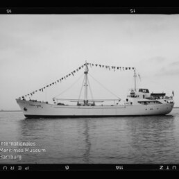 Seitenansicht eines weißen Schiffes mit zentralem Mast, an dem eine Girlande aus Signalfahnen hängt; das Boot liegt auf ruhigem Wasser vor flachem Horizont, im Foto sind Filmrand und ein Museumslogo zu sehen.