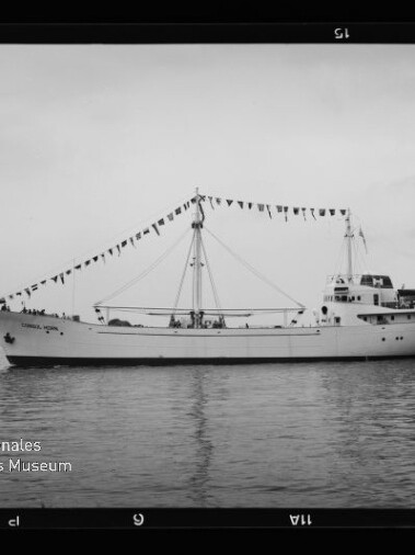 Seitenansicht eines weißen Schiffes mit zentralem Mast, an dem eine Girlande aus Signalfahnen hängt; das Boot liegt auf ruhigem Wasser vor flachem Horizont, im Foto sind Filmrand und ein Museumslogo zu sehen.
