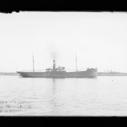 Auf dem Foto ein schlankes Dampfschiff mit einem rauchenden Schornstein und zwei Masten, das auf ruhigem Wasser vor einem flachen Küstenhorizont fährt; unten links ein Wasserzeichen des Internationalen Maritimen Museums Hamburg.