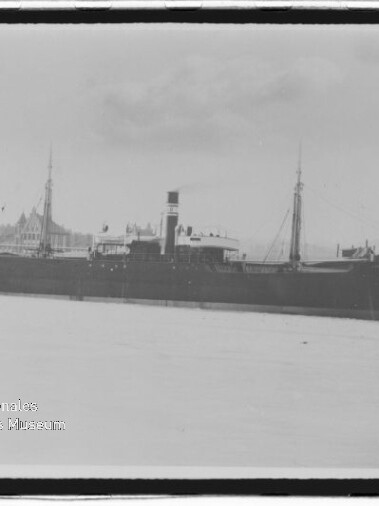 Schwarz-weiß-Foto eines großen Dampfschiffs mit Schornstein und Masten, am Kai festgemacht vor einer Stadtsilhouette mit spitzen Gebäuden, im Vordergrund Wasser und unten links ein Wasserzeichen des Internationalen Maritimen Museums Hamburg.