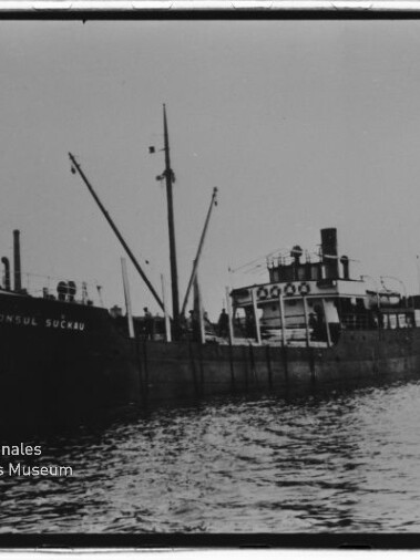 Schwarzweißaufnahme eines im Hafen liegenden Frachters mit Ladebäumen und Schornstein, auf dessen Bug der Name 'KONSUL SUCKAU' zu lesen ist; im Vordergrund Wasser, unten links das Wasserzeichen des Internationalen Maritimen Museums Hamburg.