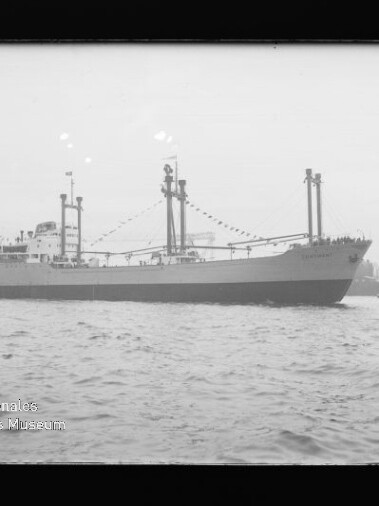 Schwarzweiß-Seitenansicht eines Frachtschiffs mit mehreren Ladebäumen und aufgezogenen Signalfahnen, auf dem Wasser vor Kaimauern und Hafenkränen, mit Wasser im Vordergrund und Museumswasserzeichen unten links.