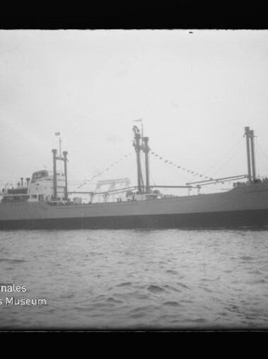 Schwarzweiß-Aufnahme eines Frachtschiffs in Seitenansicht auf ruhiger See mit mehreren Ladebäumen und Girlanden an den Masten; unten links ist das Logo des Internationalen Maritimen Museums Hamburg sichtbar.