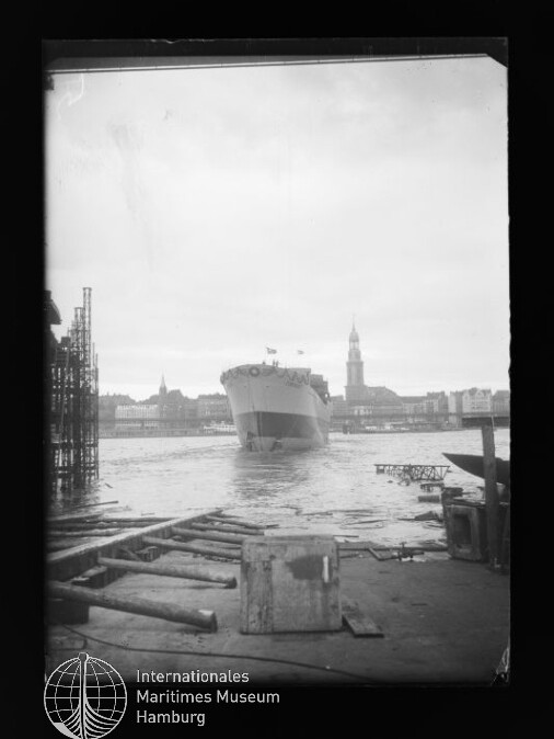 Großes Passagierschiff fährt in einen Hafen; im Vordergrund Kaianlage mit Holzbohlen und einer Kiste, im Hintergrund die Hamburger Stadtsilhouette mit dem Turm der St.-Michaelis-Kirche.