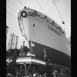 Bug des Schiffes mit dem Namen Continent auf der Werft, auf der Slipanlage aufgerichtet und mit Kranz sowie Girlanden geschmückt, vor Kränen und Gerüsten und einem dicht gedrängten Publikum im Vordergrund.