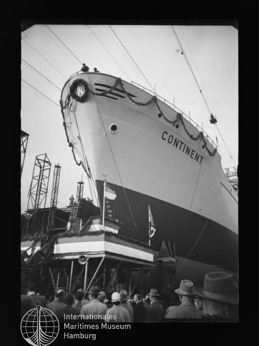Bug des Schiffes mit dem Namen Continent auf der Werft, auf der Slipanlage aufgerichtet und mit Kranz sowie Girlanden geschmückt, vor Kränen und Gerüsten und einem dicht gedrängten Publikum im Vordergrund.