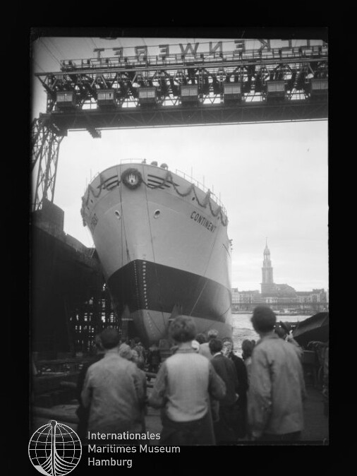 Bugansicht des Schiffs CONTINENT auf einer Werft-Slipanlage während des Stapellaufs, mehrere Zuschauer im Vordergrund, ein hohes Portalkran-Gerüst mit Schriftzug darüber und ein Kirchturm am gegenüberliegenden Ufer im Hintergrund.