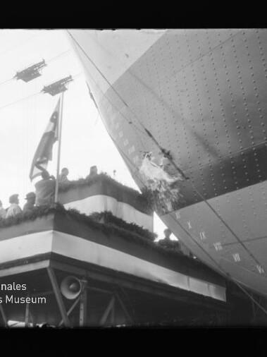 Schwarz-weiß-Aufnahme: Menschen auf einem geschmückten Tribünenpodium mit Flagge beobachten ein großes Luftschiff, dessen Außenhülle rechts deutlich einen Riss/Loch und Nieten zeigt.