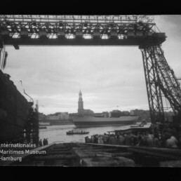 Schwarzweißfoto, durch das Stahlgerüst eines großen Auslegerkrans gerahmter Blick auf ein großes Schiff im Hafen, vor dem Ufer eine Menschenmenge und im Hintergrund eine Stadtsilhouette mit hohem Kirchturm; linke untere Ecke zeigt das Logo des Internationalen Maritimen Museums Hamburg.