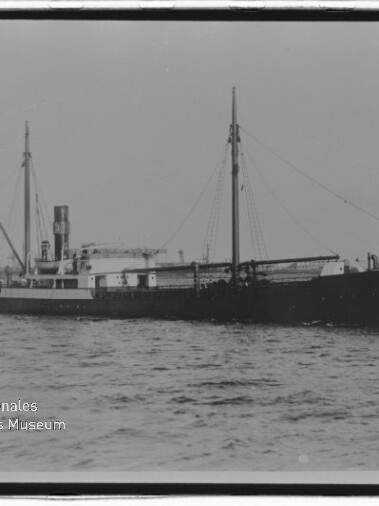 Schwarzweißaufnahme eines historischen Frachtschiffs in Seitenansicht auf ruhiger See, mit prominentem Schornstein, zwei Masten und Ladebaum; unten links sichtbar das Logo des Internationalen Maritimen Museums Hamburg.