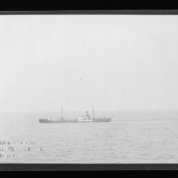 Schwarzweiß-Foto eines kleinen Frachtdampfers mittig am Horizont auf ruhiger See unter weitem Himmel, im unteren linken Bildbereich ein Wasserzeichen des Internationalen Maritimen Museums Hamburg.