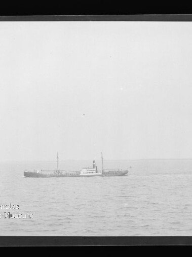 Schwarzweiß-Foto eines kleinen Frachtdampfers mittig am Horizont auf ruhiger See unter weitem Himmel, im unteren linken Bildbereich ein Wasserzeichen des Internationalen Maritimen Museums Hamburg.