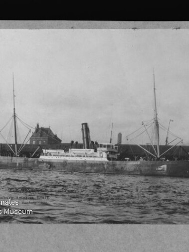 Schwarz-weiß-Fotografie eines großen Dampfschiffs mit zentralem Schornstein und zwei Masten sowie Ladebäumen, längsseits am Kai vor historischen Gebäuden, welliges Wasser im Vordergrund und Museumswasserzeichen unten links.