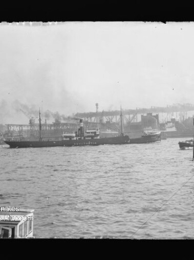 Schwarz-weiß-Aufnahme eines Frachtschiffs auf ruhigem Hafenwasser vor industriellen Kaianlagen mit Kränen, Lagerhallen und rauchenden Schornsteinen, im Vordergrund Wellen und Poller sowie ein Museumswasserzeichen unten links.