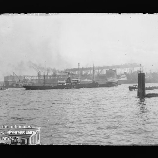 Schwarz-weiß-Aufnahme eines Frachtschiffs auf ruhigem Hafenwasser vor industriellen Kaianlagen mit Kränen, Lagerhallen und rauchenden Schornsteinen, im Vordergrund Wellen und Poller sowie ein Museumswasserzeichen unten links.