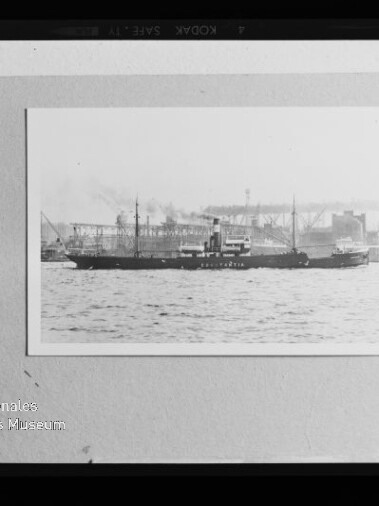Gerahmtes Schwarzweißfoto eines dampfgetriebenen Frachtschiffs auf dem Wasser vor einer Industriehafen-Kulisse, auf hellem Karton montiert und mit dem Stempel 'Internationales Maritimes Museum Hamburg' unten links.