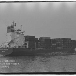 Schwarzweiß-Foto eines Containerschiffs (Contship Barcelona) mit gestapelten Containern, das auf ruhiger See fährt, mit Wasserzeichen des Internationalen Maritimen Museums Hamburg unten links.