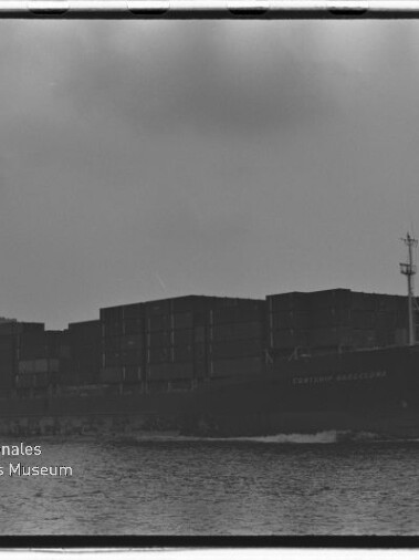 Schwarzweißfoto eines beladenen Containerschiffs in Seitenansicht, das mit mehreren Containerstapeln über ruhigem Wasser fährt; unten links ist das Logo und der Schriftzug des Internationalen Maritimen Museums Hamburg zu sehen.