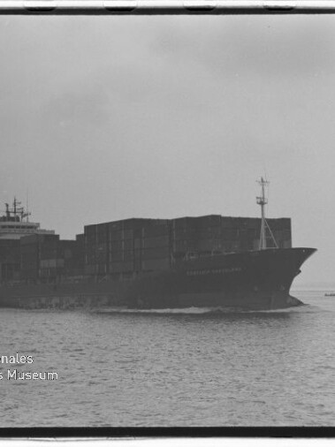 Schwarzweißaufnahme eines beladenen Containerschiffs mit gestapelten Containerkästen, das bei ruhiger See vor bedecktem Himmel fährt; rechts im Hintergrund eine kleinere Plattform und links unten das Logo des Internationalen Maritimen Museums Hamburg.