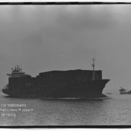 Großes beladenes Containerschiff als dunkle Silhouette auf ruhiger See, rechts ein kleines Begleitboot vor wolkigem Himmel; unten links das Logo des Internationalen Maritimen Museums Hamburg.