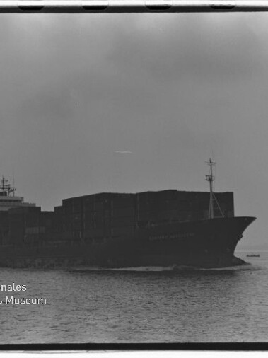 Großes beladenes Containerschiff als dunkle Silhouette auf ruhiger See, rechts ein kleines Begleitboot vor wolkigem Himmel; unten links das Logo des Internationalen Maritimen Museums Hamburg.