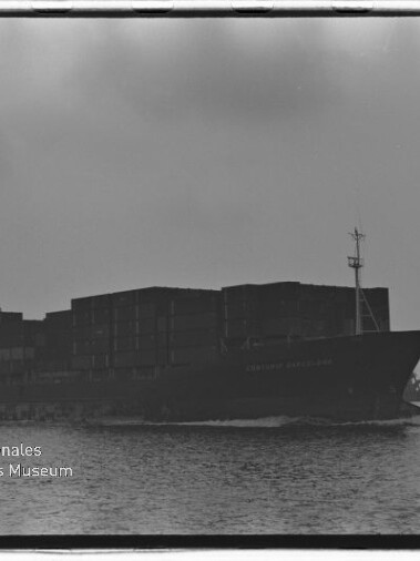 Schwarzweißaufnahme eines großen Containerschiffs in Seitenansicht auf ruhigem Wasser, mit aufgetürmten Containerstapeln und dem Logo/Schriftzug des Internationalen Maritimen Museums Hamburg unten links.