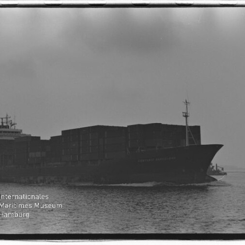 Schwarzweißaufnahme eines großen Containerschiffs in Seitenansicht auf ruhigem Wasser, mit aufgetürmten Containerstapeln und dem Logo/Schriftzug des Internationalen Maritimen Museums Hamburg unten links.