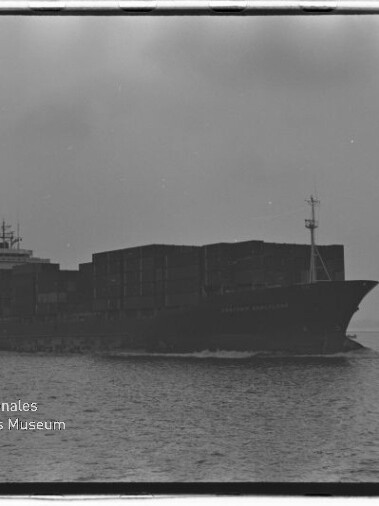 Schwarz-weiß-Foto eines großen, beladenen Containerschiffs, das bei ruhigem Wasser und bedecktem Himmel fährt und rechts von einem kleinen Schlepper begleitet wird; unten links sichtbares Wasserzeichen des Internationalen Maritimen Museums Hamburg.