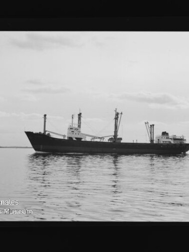 Schwarzweißfoto eines Frachtschiffs in Seitenansicht auf ruhiger See unter leicht bewölktem Himmel, mit Museumswasserzeichen unten links.