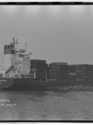 Schwarzweiß-Foto eines beladenen Containerfrachters auf ruhiger See, seitliche Ansicht mit aufgetürmten Containern und dem Logo des Internationalen Maritimen Museums Hamburg unten links.