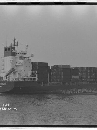 Schwarzweiß-Foto eines beladenen Containerfrachters auf ruhiger See, seitliche Ansicht mit aufgetürmten Containern und dem Logo des Internationalen Maritimen Museums Hamburg unten links.