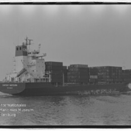 Schwarzweiß-Foto eines beladenen Containerschiffs auf ruhiger See mit mehreren gestapelten Seecontainern, sichtbarem Filmrahmen und Wasserzeichen des Internationalen Maritimen Museums Hamburg unten links.